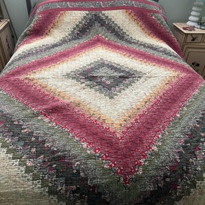 May include: A quilt featuring a large diamond-shaped pattern composed of small, multi-colored fabric squares. The design incorporates shades of deep red, olive green, cream, and muted floral prints, creating a concentric, geometric layout.