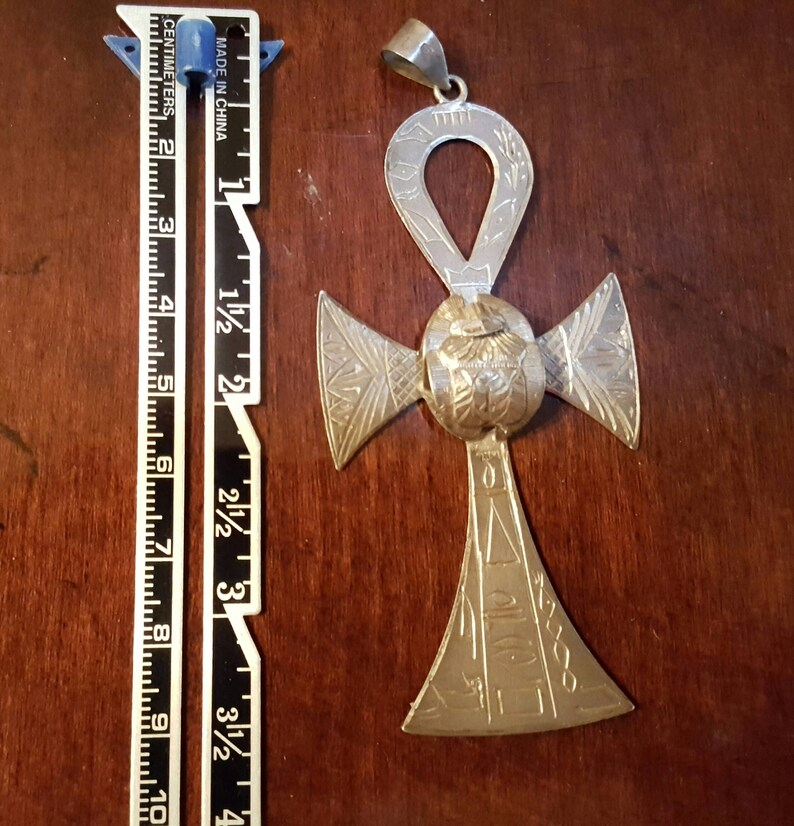 May include: Silver Ankh pendant with intricate details and a loop at the top for hanging. The pendant is about 3 inches tall and features a cross shape with a circle in the center.