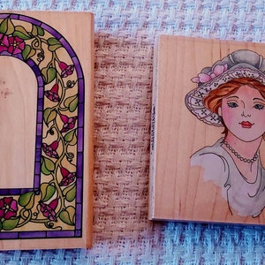 May include: Two wooden stamps, one with a stained glass arch design featuring purple, green, and yellow colors, and the other with a woman's face wearing a hat with pink flowers and a pearl necklace.
