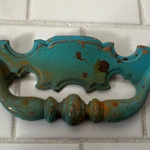 May include: A distressed turquoise drawer pull with a curved handle and ornate detailing. The metal hardware has a weathered appearance with hints of gold and rust, suggesting an antique or vintage style.