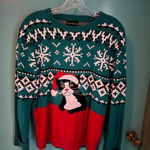 May include: A green Christmas sweater with a pattern of white snowflakes and black accents. The bottom half is red, with a black and white cat wearing a Santa hat. The sweater is hanging on a clear hanger.