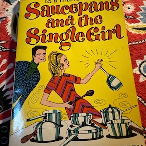 May include: A vintage cookbook titled "Saucepans and the Single Girl" with a yellow cover. The cover art features a cartoon illustration of a woman cooking, a man, and several pots and pans. The book's title and author's names are in bold, red text.