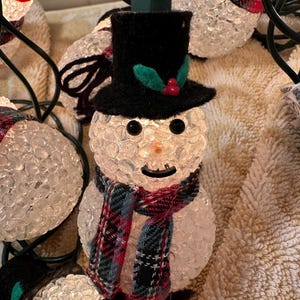 May include: A string of clear, bubble-textured snowman lights with black hats, red berries, and plaid scarves. The lights are strung together with green wire.
