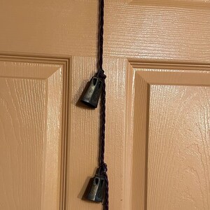 May include: A set of three small, antique-looking bells hanging from a dark purple cord. The bells are made of metal and have a slightly aged appearance. The cord is tied to a hook on a light brown wooden door.