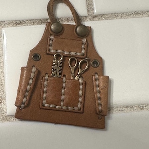 May include: A miniature brown leather apron with white stitching, a loop for hanging, and two pockets. The pockets hold tiny metal scissors and a ruler. The apron has metal rivets and is designed to look like a miniature apron.