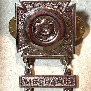May include: A silver-colored mechanic badge with a cross-shaped design and a central gear emblem. The word "MECHANIC" is displayed on a rectangular bar below the cross. Two gold-colored pin backs are visible on the sides.