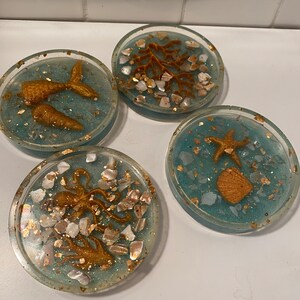 May include: Four round blue and gold resin coasters with different sea-themed designs. Each coaster features a different sea creature, including a mermaid tail, a starfish, an octopus, and a coral branch. The coasters are decorated with gold glitter and white seashells.