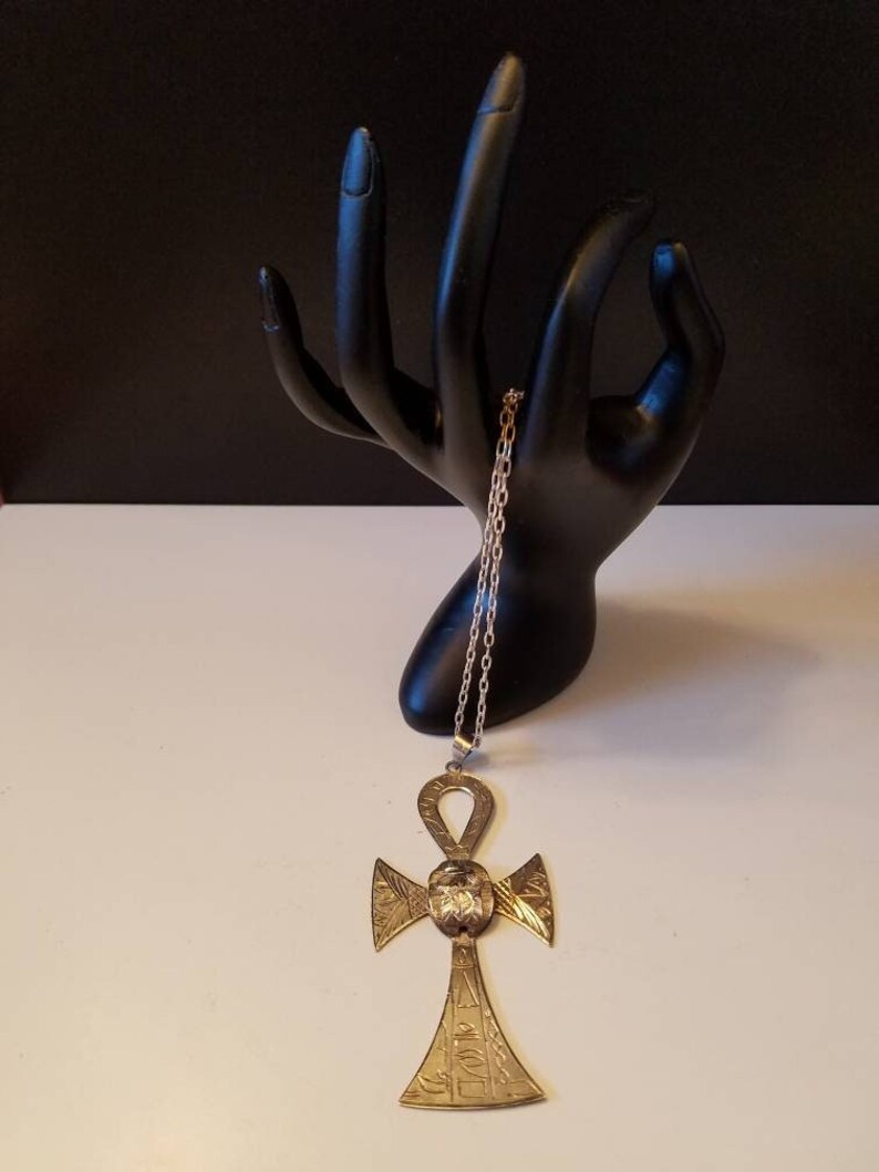 May include: A gold-toned Ankh pendant necklace with intricate details and an engraved design. The pendant is hanging from a silver chain.