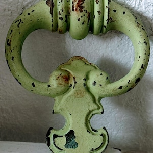 May include: A distressed, light green metal handle with an oval grip and decorative base. The handle shows signs of wear with exposed rust and black speckling, suggesting an antique or vintage aesthetic. The handle is mounted on a white surface.