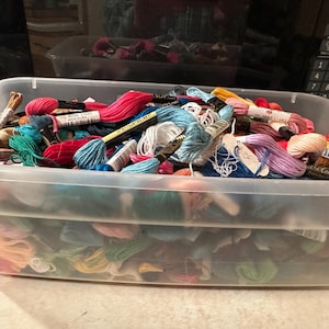 May include: A clear plastic storage bin filled with colorful embroidery floss. The floss is in various shades of pink, blue, green, and white. Some floss has tags attached. The bin is sitting on a light-colored surface.