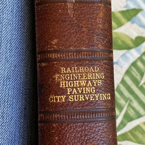 May include: A brown leather-bound book with gold lettering. The title is "I.C.S. Reference Library". The book lists the subjects covered: "Railroad Engineering, Highways, Paving, City Surveying". The bottom of the book reads "133 International Correspondence Schools".