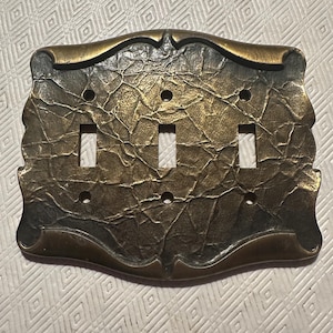 May include: Ornate bronze triple light switch cover with a textured, aged appearance. The cover has three rectangular switch openings and decorative edges. The finish has a mix of dark and light bronze tones.