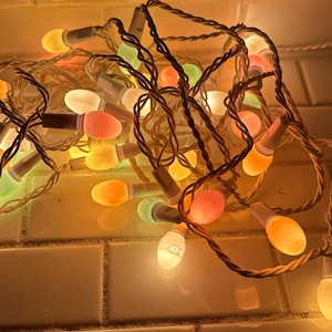 May include: A string of colorful, egg-shaped Christmas lights. The lights are a mix of orange, pink, yellow, and green, with white bases. The cord is a twisted white and brown. The lights are on a white tiled surface.