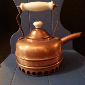 May include: A copper tea kettle with a white handle and knob. The kettle has a round base with several small holes. The handle and knob are white. The kettle is sitting on a blue chair.