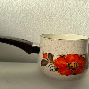 May include: A vintage small saucepan with a brown handle and a floral design. The cream-colored pot features a large orange flower with brown leaves and accents. The handle is dark brown, and the interior is cream-colored.
