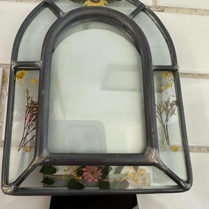 May include: An arched glass frame with a dark metal border, featuring pressed dried flowers including a yellow sunflower at the top, small yellow sprigs on the sides, and a pink clover-like flower at the base, mounted on a black stand.
