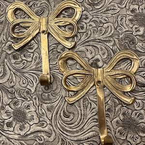 May include: Two gold-colored metal wall hooks in a bow shape. The hooks have a decorative design and a curved hook at the bottom. The background is a textured, silver-colored surface with a floral pattern.