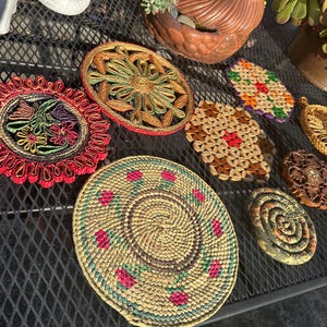 May include: A collection of handmade coasters in various shapes and designs. The coasters feature woven and braided textures in a variety of colors, including red, green, yellow, and brown. Some have floral patterns, while others have geometric designs.