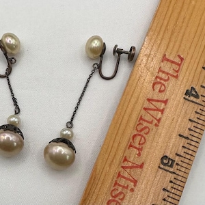 May include: A pair of vintage clip-on earrings. Each earring features a cream-colored pearl at the top, connected by a chain to a smaller pearl and a larger, pearlescent drop. The metal components have a dark, aged finish.