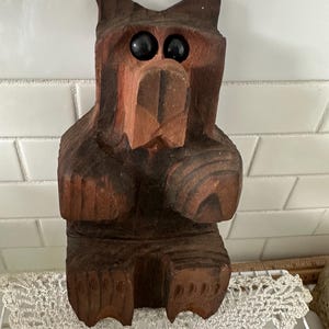 May include: A hand-carved wooden bear figurine with dark brown and reddish-brown tones. The bear has large, black eyes and is seated with its paws clasped. The carving details the bear's fur and facial features, creating a rustic, folk art aesthetic.
