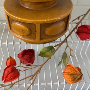 May include: A vintage, two-tiered, wooden candle holder in shades of brown and tan. The candle holder is decorated with oval-shaped details. A branch with red and orange flowers and green leaves is in the foreground.