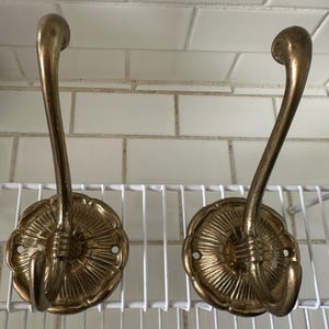 May include: Two antique brass wall hooks. Each hook features a decorative floral base and a curved, elongated hook. The hooks are mounted on a white wire shelf against a backdrop of white tiles. The brass has a warm, aged patina.