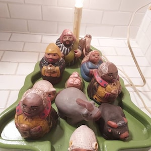 May include: A green ceramic Christmas tree with a light bulb on top, surrounded by figurines of the nativity scene, including Mary, Joseph, baby Jesus, a donkey, a cow, and sheep. The figurines are painted in various colors, including brown, white, yellow, and blue.