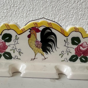 May include: A white ceramic utensil holder with a rooster design. The rooster is yellow, red, and black with a green and yellow floral pattern around it. The holder has five slots for utensils.