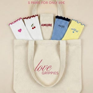 Könnte beinhalten: Eine cremefarbene Tragetasche aus Segeltuch mit fünf Paar Socken. Die Socken sind weiß, blau und gelb, mit Rüschenkanten und verschiedenen Designs, darunter ein Herz, Kirschen und die Wörter "AMORE", "stargirl" und "wild at". Auf der Tragetasche steht "love GRIPPIES".