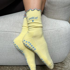 Könnte beinhalten: Ein Paar gelbe Socken mit blauen Akzenten. Die Socken haben einen gerüschten oberen Rand und die Worte "wild at" sind in Blau gestickt, zusammen mit einem kleinen blauen Herz. Die Sohlen haben blaue herzförmige Griffe.