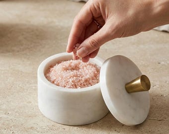 May include: A cylindrical white marble salt cellar filled with coarse pink Himalayan salt. The container features a matching circular marble lid topped with a small, conical brass knob, providing a functional and elegant kitchen accessory.