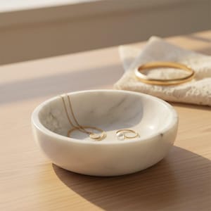 Puede incluir: Un cuenco redondo de m&aacute;rmol blanco con vetas grises que contiene un collar de cadena dorada fina con un peque&ntilde;o colgante circular y un anillo dorado con una perla blanca. Un brazalete dorado descansa sobre un trozo de encaje blanco.