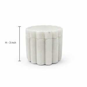 May include: A white marble container with a scalloped, fluted exterior design and a matching lid. The cylindrical vessel features a vertical ribbed pattern around its circumference. A measurement indicator shows a height of 3 inches.