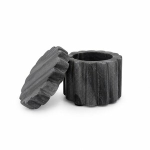 May include: A dark gray stone container with a matching circular lid. The cylindrical base and the lid both feature a distinct vertical fluted or ribbed texture around their outer edges. The lid is positioned upright next to the open container.