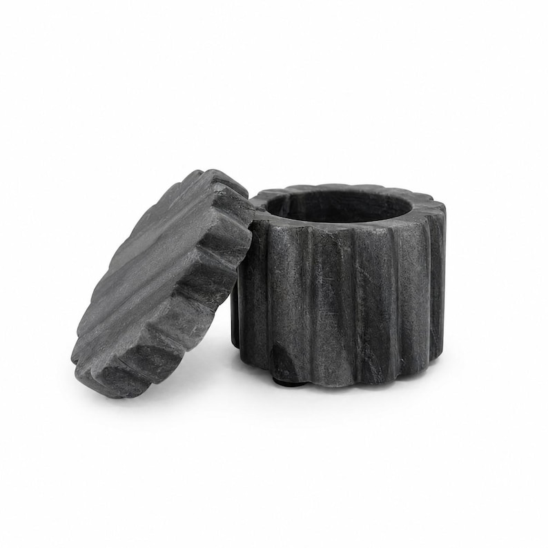 May include: A dark gray stone container with a matching circular lid. The cylindrical base and the lid both feature a distinct vertical fluted or ribbed texture around their outer edges. The lid is positioned upright next to the open container.