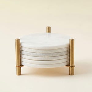 May include: A stack of six circular white marble coasters held within a vertical brass metal stand. The stand features three thin cylindrical posts connected by a base frame, securely containing the smooth, polished stone discs in a neat pile.
