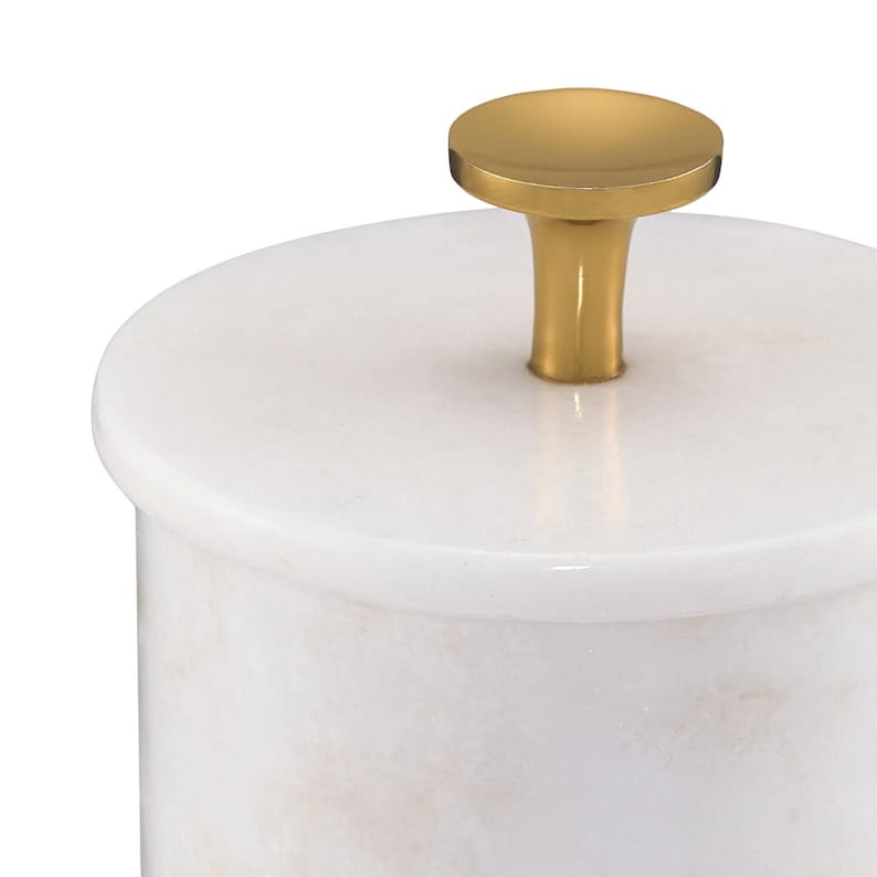 May include: A cylindrical white marble container with a smooth, polished surface and a circular lid. The lid features a centered, solid gold-toned metal knob with a flared base and a flat, circular top, providing a contrast to the stone.