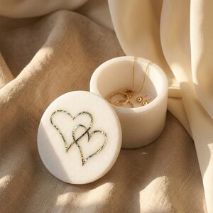 May include: A round white marble trinket box with a removable lid featuring an inlaid iridescent shell design of two interlocking hearts. The open container holds a thin gold chain necklace and a pair of small gold stud earrings inside.