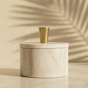 May include: A cylindrical beige stone container with a smooth, polished finish and a matching circular lid. A small, tapered metallic gold knob is centered on top of the lid, providing a contrasting accent to the neutral, textured stone surface.