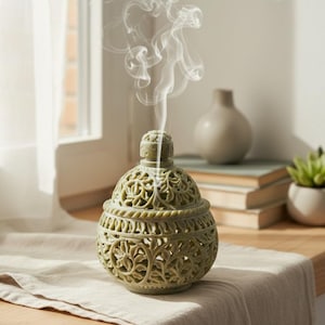 May include: A green, intricately carved incense burner with a domed lid. The burner features a floral design and is emitting a wisp of smoke. The burner is sitting on a beige cloth, with a window and other decorative items in the background.