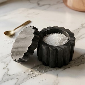 Puede incluir: Un salero de mármol gris oscuro con un exterior festoneado y estriado, junto con una tapa extraíble a juego. El recipiente está lleno de cristales de sal marina blanca gruesa y una pequeña cuchara de metal dorado descansa cerca.