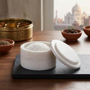 May include: A white marble salt cellar with a matching circular lid, filled with coarse white salt crystals. The container sits on a rectangular black slate board, accompanied by small terracotta bowls and a large brass spice container.