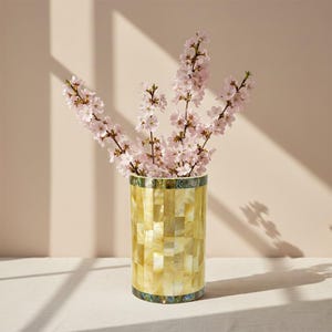Op de afbeelding: Een cilindervormige vaas met een mozaïekpatroon in tinten geel en goud, met takken van roze kersenbloesems. De vaas heeft een decoratieve band aan de boven- en onderkant. De achtergrond is zacht beige.