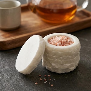 May include: A cylindrical white marble salt cellar with a rough, chiseled exterior texture and a matching circular lid. The container is filled with coarse pink Himalayan salt crystals, with a few loose salt grains scattered on the surface.