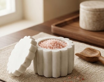 May include: A white marble salt cellar with a scalloped, fluted exterior design and a matching removable lid. The container is filled with coarse pink Himalayan salt crystals, and a small wooden scoop is positioned nearby on a textured mat.