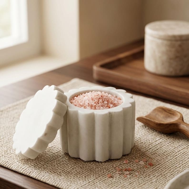 May include: A white marble salt cellar with a scalloped, fluted exterior design and a matching removable lid. The container is filled with coarse pink Himalayan salt crystals, and a small wooden scoop is positioned next to the vessel.