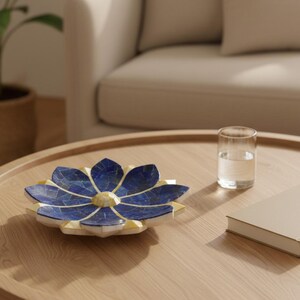 Op de afbeelding: Een decoratieve, bloemvormige schaal met een blauw en geel mozaïektegelontwerp. De schaal staat op een houten tafel, naast een glas water en een gesloten boek. De bloemblaadjes van de schaal zijn diepblauw, met gele accenten.