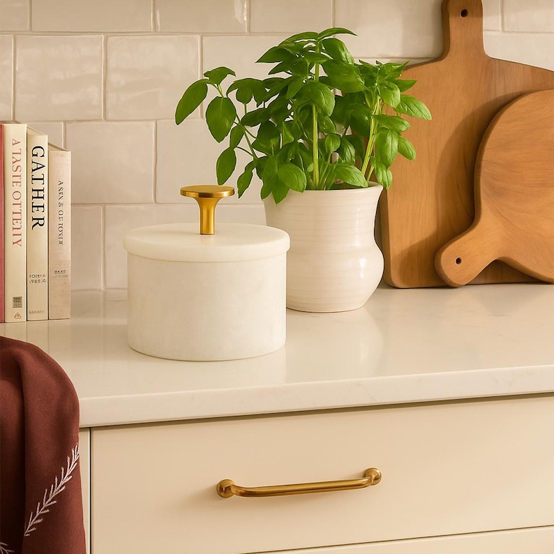 May include: A cylindrical white marble container with a gold-toned metal knob handle on the lid. The container sits on a white surface next to a white ceramic pot containing a green basil plant, two wooden cutting boards, and three books.