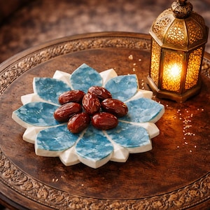Op de afbeelding: Een houten dienblad met bloemmotief draagt een blauw-witte keramische schaal met dadels. Rechts staat een gouden lantaarn met gedetailleerde uitsparingen, die een warm licht uitstraalt. De afbeelding roept een Midden-Oosterse sfeer op.
