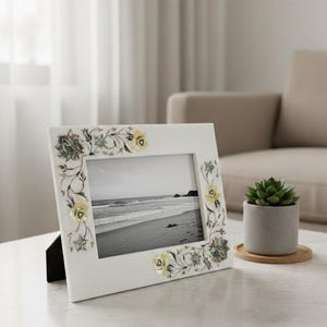 Könnte beinhalten: Ein weißer Bilderrahmen mit floralen Einlegearbeiten. Er enthält ein Schwarz-Weiß-Foto einer Strandszene. Das florale Design umfasst gelbe, grüne und irisierende Akzente. Der Rahmen steht auf einem weißen Tisch.